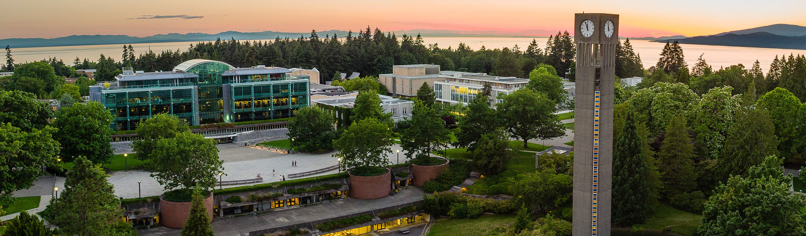 Aerial photo of the UBC Vancouver campus with a clock tower in the foreground with buildings, mountains, and oceans in the background at sunset.