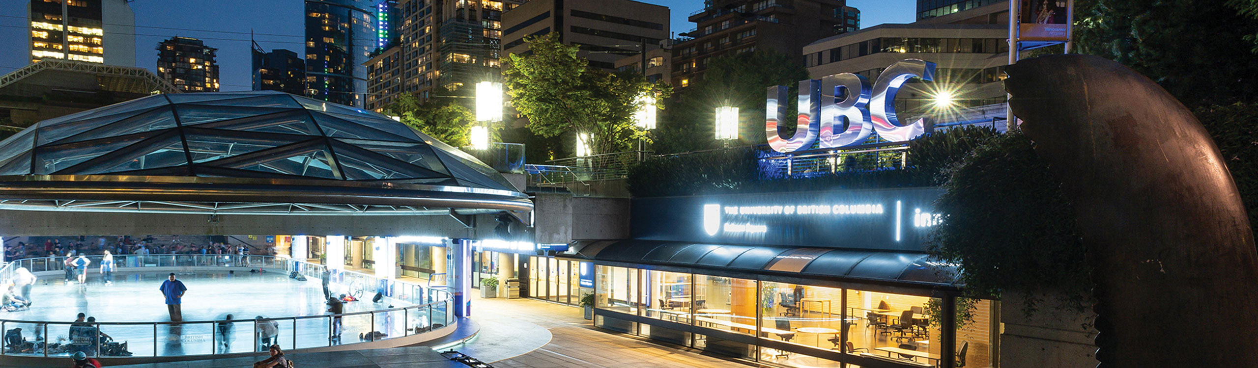 Large metallic "UBC" sign and covered skating rink at the UBC Robson Square site in downtown Vancouver in the evening.